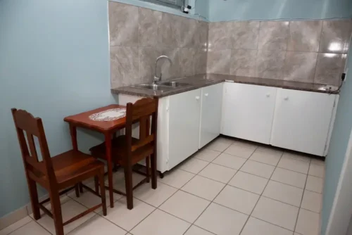 Kitchenette corner in the self-catering room with white cupboards, a sink and a small wooden dining table for two.