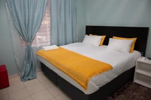 The Single Room bed with a yellow throw, a white bedside table and blue curtains.