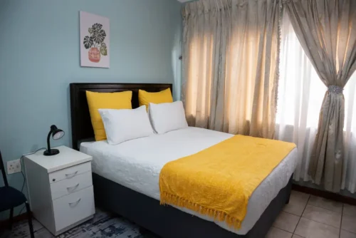 Bed with yellow cushions, a white bedside table and a chair in the Single Room.
