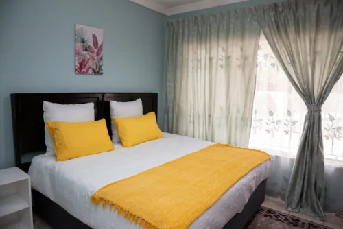 Bed with white linen, a yellow throw and yellow cushions in the Single Room, next to a window with sheer curtains.
