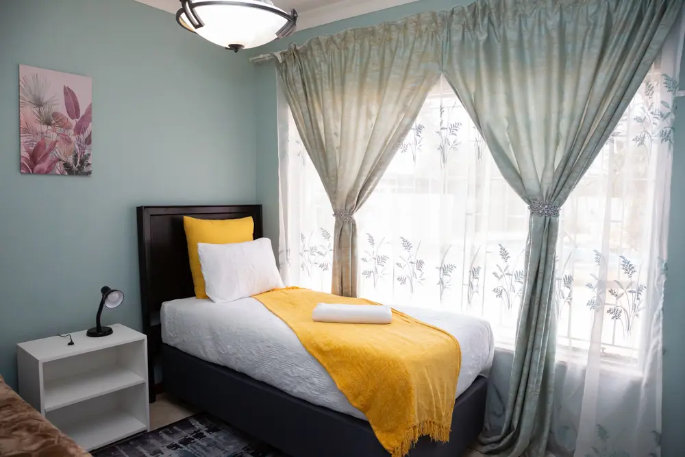 Bed with a yellow throw beside a large window with sheer curtains in the Deluxe Room.